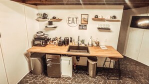 A compact kitchen with a wooden countertop, a small refrigerator, and various kitchen appliances. There are shelves with kitchenware and a wall-mounted sign that reads "EAT GOOD MOOD". - Wild And Wonderful Silos (Harpers Ferry)