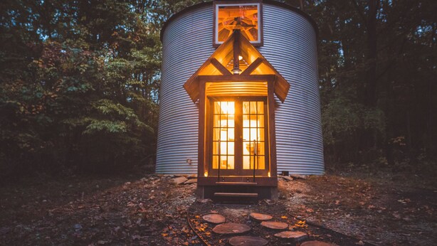 A cylindrical metal building with a lit entrance, surrounded by trees. - Wild And Wonderful Silos (Harpers Ferry)