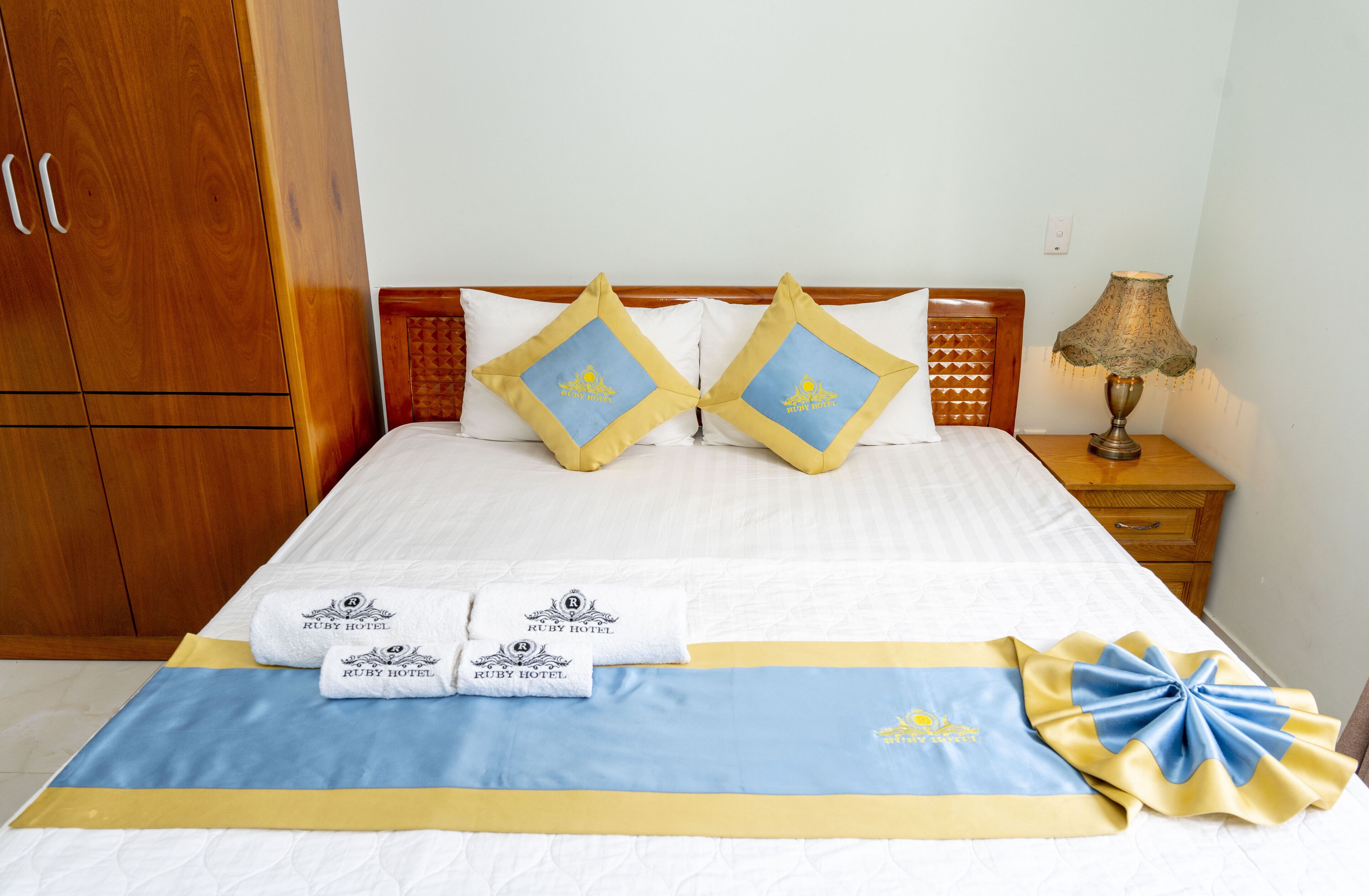 A neatly made bed with a blue and yellow bedspread, two matching pillows, and a set of towels with the Ruby Hotel logo.