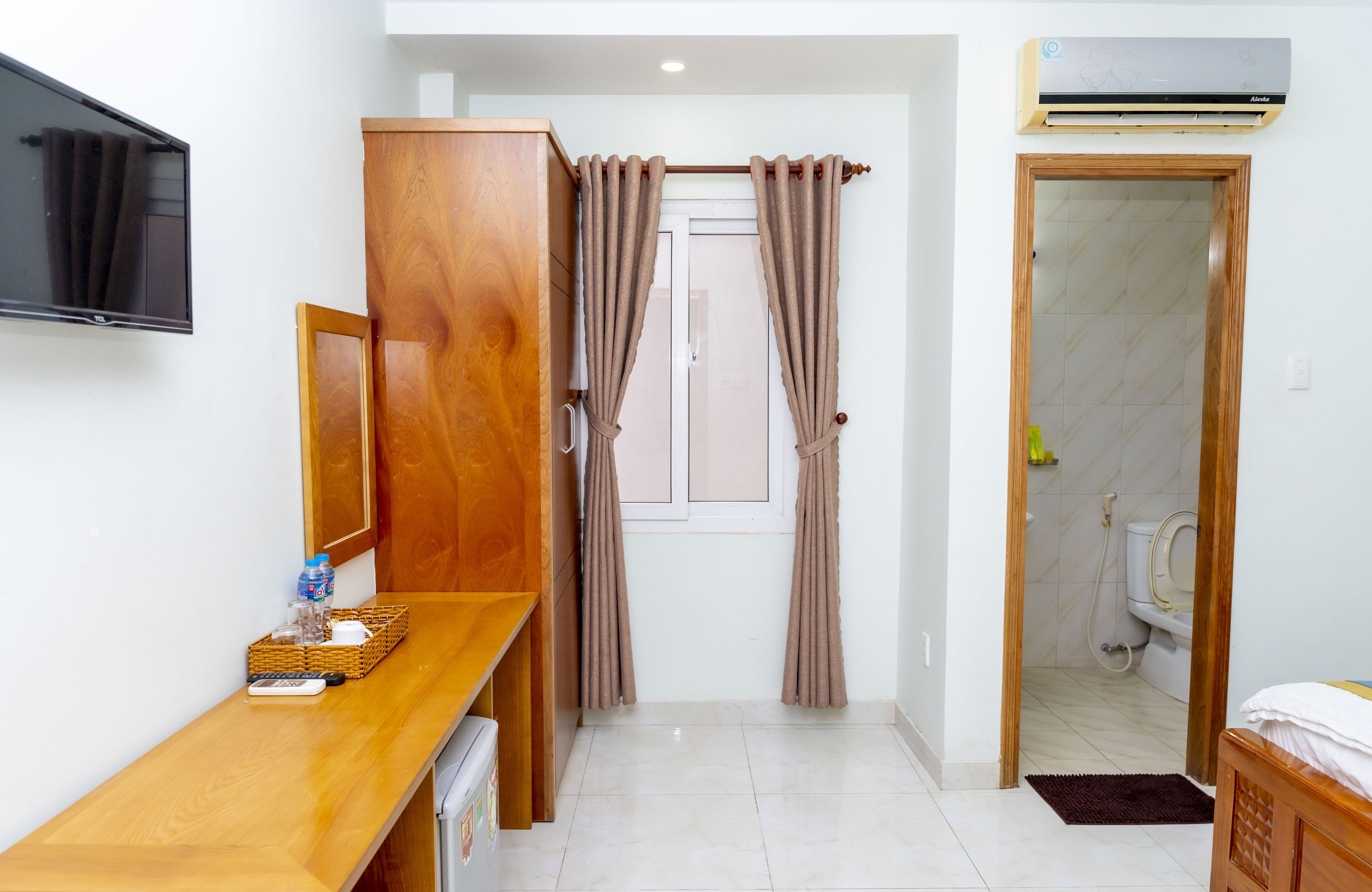 Habitación de hotel con escritorio de madera, televisor, armario, baño con inodoro y ducha, y cortinas color beige.