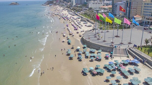 Een strand met parasols en mensen, een promenade met vlaggen, en gebouwen op de achtergrond.
