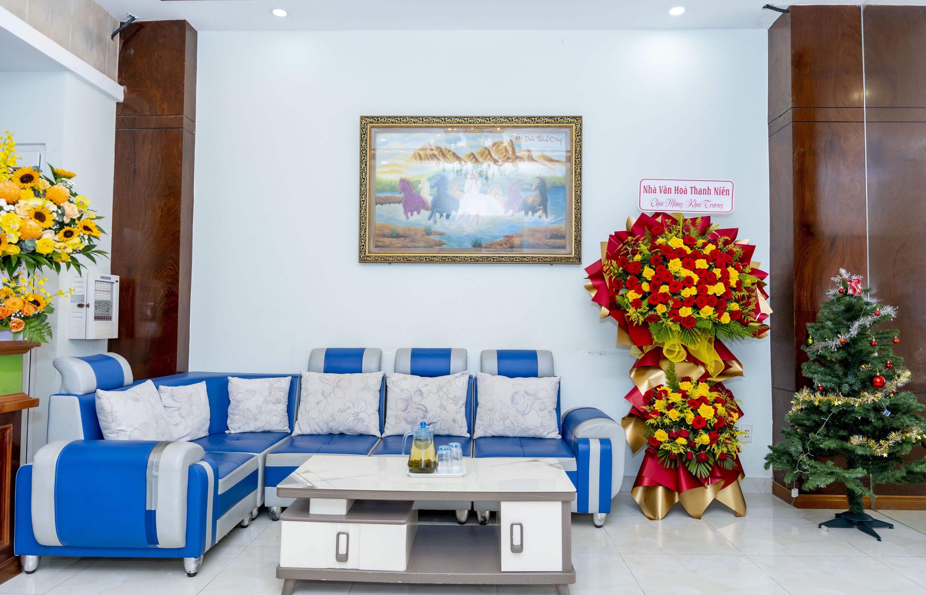 Un espace d’accueil avec un ensemble de canapés bleu et blanc, une table basse, un tableau encadré, un grand arrangement floral et un sapin de Noël.