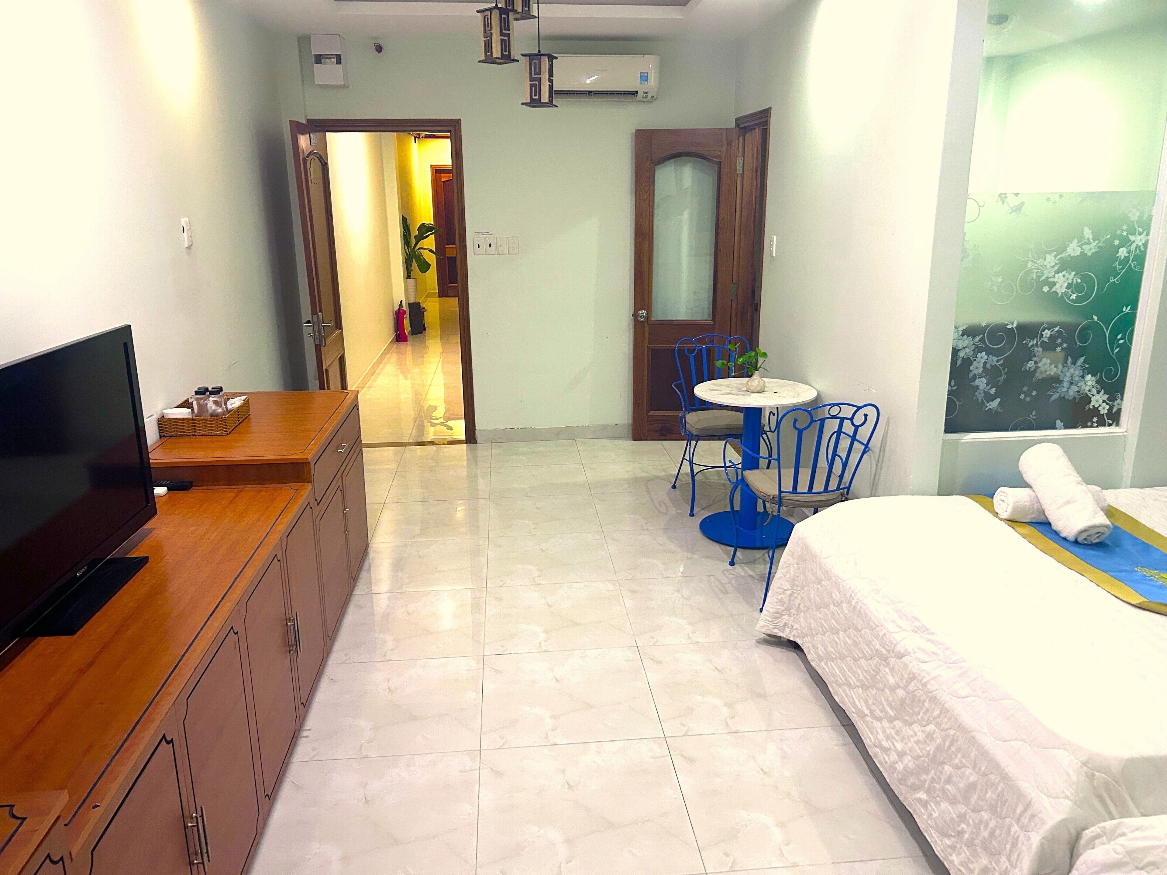 A hotel room with a bed, a TV, a desk, a chair, and a small table. There is a door leading to a hallway with a plant and a fire extinguisher.
