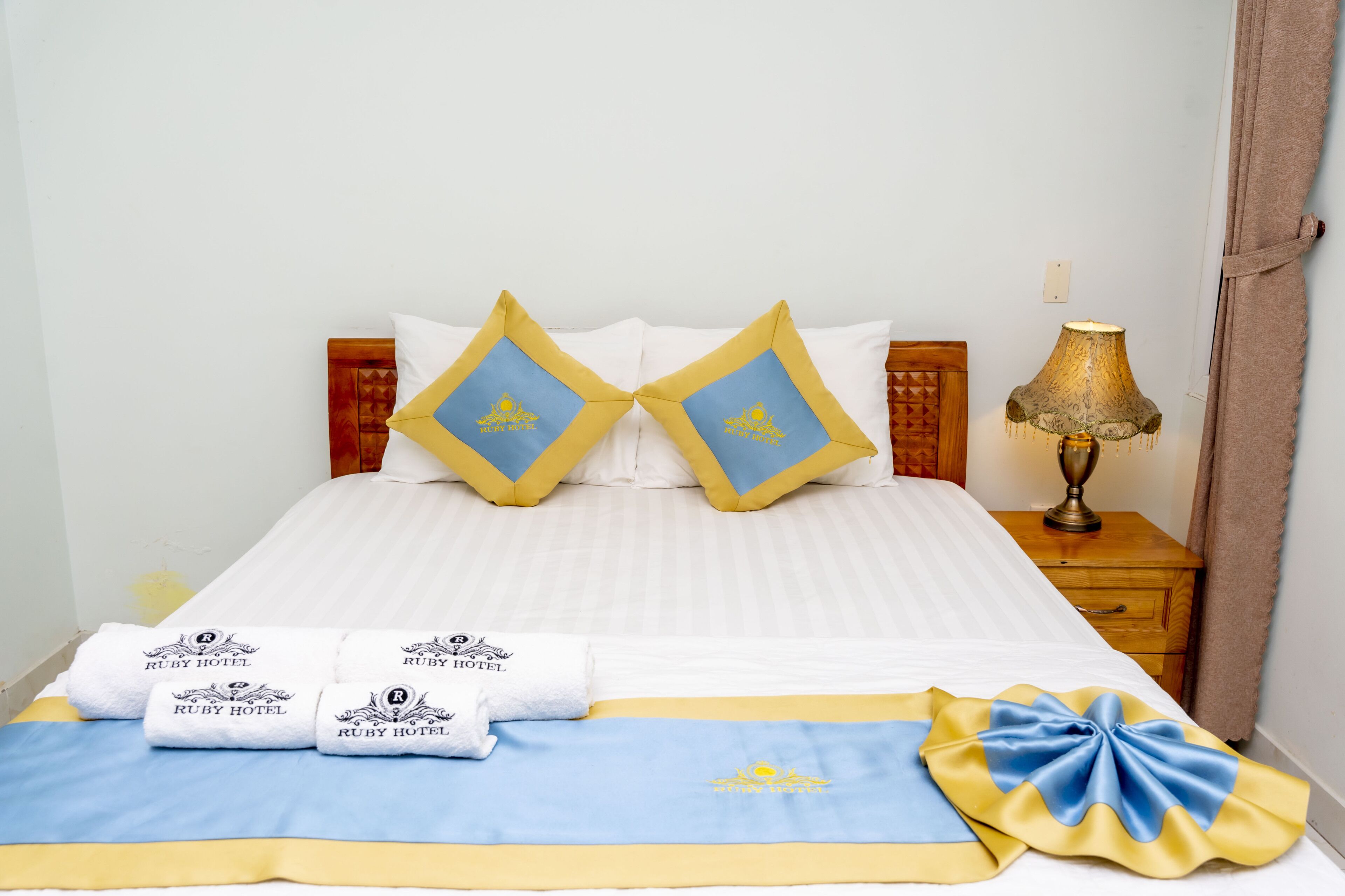 A neatly made bed with a blue and yellow bedspread, two matching pillows, and two towels with the hotel's logo.