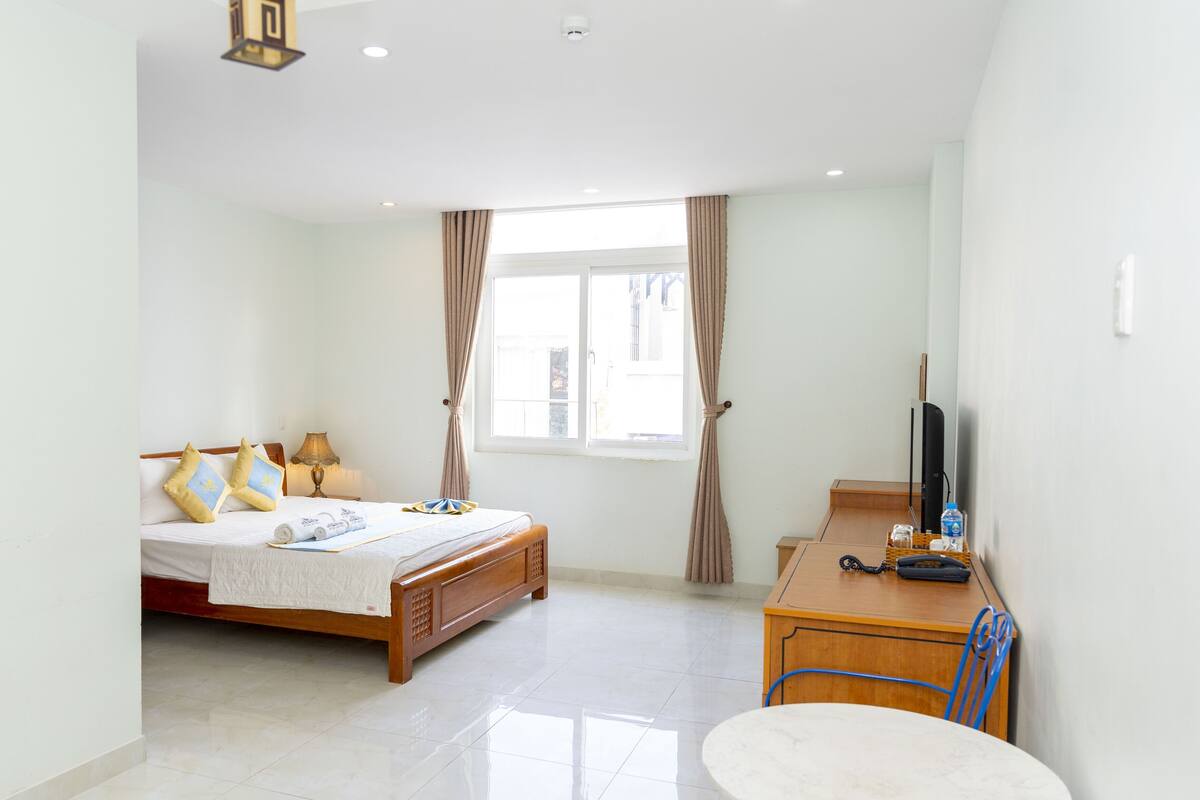 A bedroom with a bed, a desk, a chair, a TV, and a window with curtains.