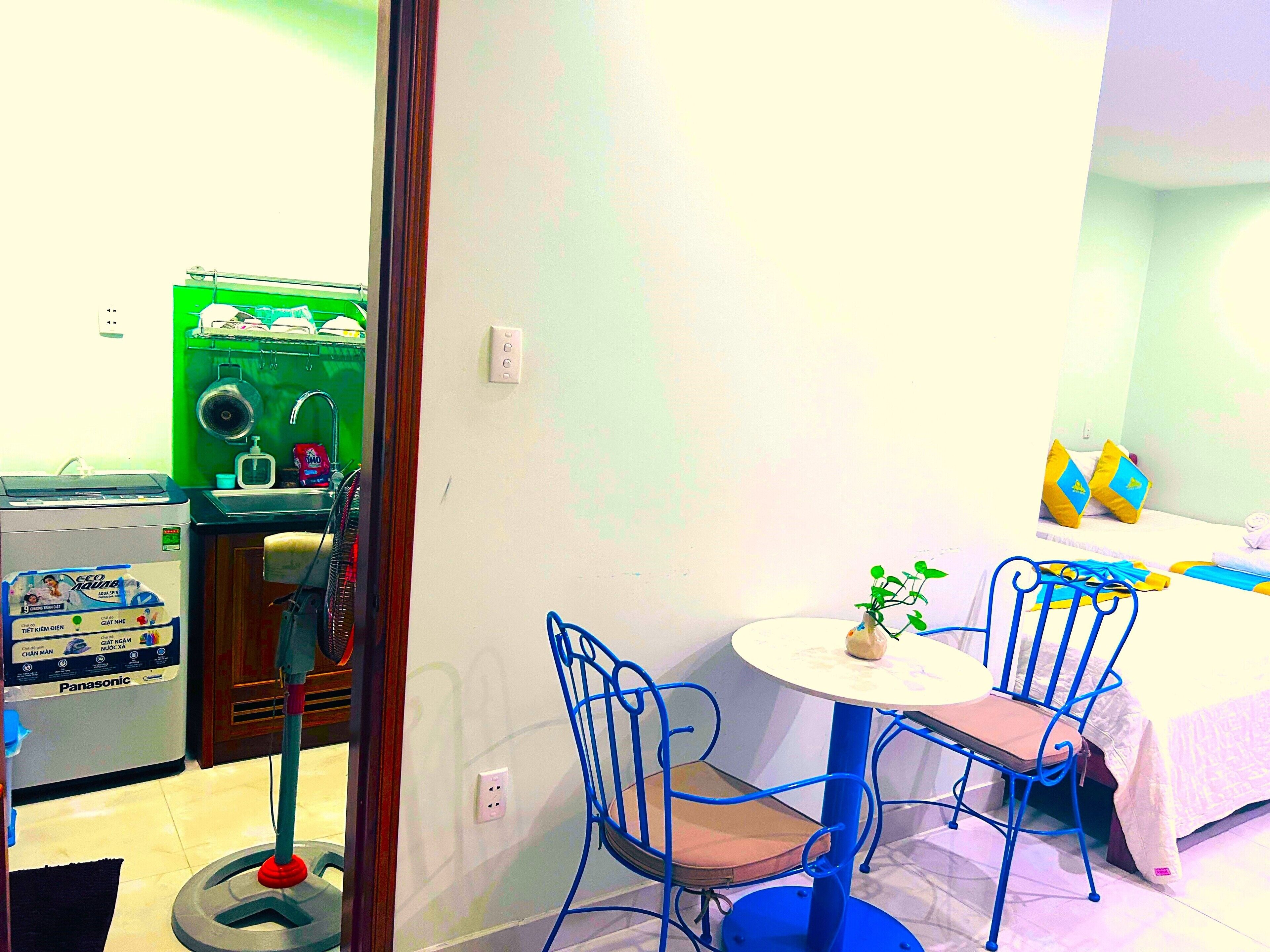 A compact room with a kitchenette, a dining area with blue chairs, and a bed with yellow pillows.