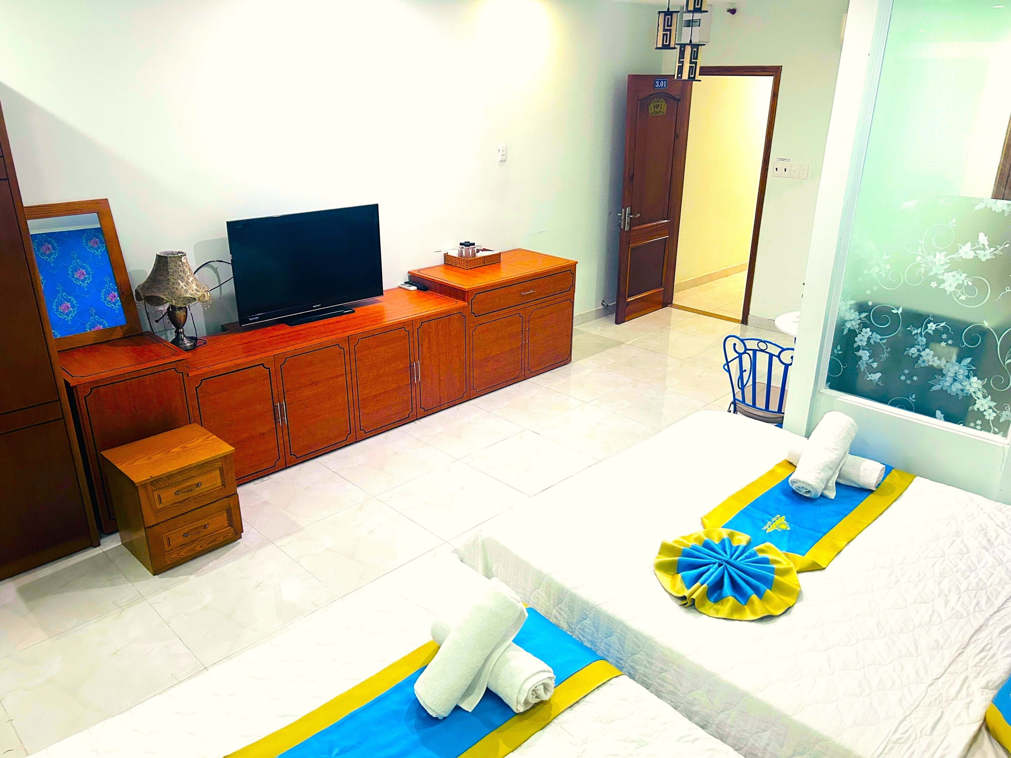 A hotel room with a wooden cabinet, a television, a small table, a bed with a blue and yellow towel, and a window with a floral view.