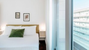 Ein Hotelzimmer mit Bett, zwei gerahmten Bildern an der Wand, einem Nachttisch mit Lampe und einem großen Fenster mit Blick auf Gebäude.
