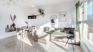 Ein modernes Wohnzimmer mit einer Eckcouch, einem Esstisch mit Stühlen, einem kleinen runden Tisch mit Lampe und einem großen Fenster mit Vorhängen.