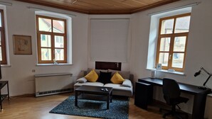 Un moderno salón con sofá, una mesita de centro, un escritorio y una computadora. Hay dos ventanas con persianas, un radiador y un marco de fotos en la pared.