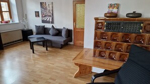 Una sala de estar con un sofá gris, una mesa de centro de madera y una silla negra. Se observa una pared con un cuadro enmarcado y una repisa decorativa con diversos objetos.