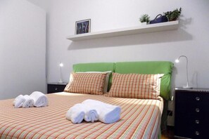 Una cama bien hecha con ropa de cama a rayas, almohadas y toallas enrolladas sobre una pared blanca.