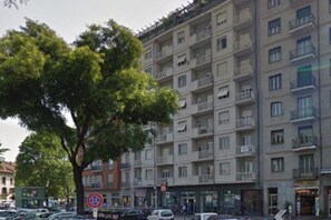Een meerlaags gebouw met balkons, een boom ervoor en geparkeerde auto's.