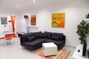 Una sala de estar moderna con un sofá seccional negro, una mesa de centro blanca y un tapete colorido. Hay un comedor con una mesa blanca y sillas rojas, y una cocina con armarios blancos.