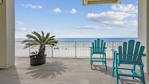 A balcony with a view of the beach, two turquoise Adirondack chairs, and a potted palm plant.