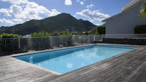 Una piscina rectangular con cubierta de madera, rodeada por una cerca blanca, con colinas verdes exuberantes y un cielo despejado de fondo.