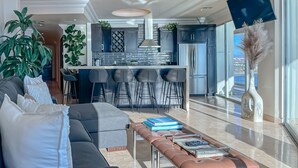 A modern living room with a gray sofa, a coffee table with books, and a view of the kitchen and balcony. - LuxuryCorner-PrivateJacuzzi-LasOlasCondo-Rosarito (Playas de Rosarito)