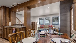 Un comedor amplio con una mesa de madera dispuesta para una comida, una cocina con armarios de madera, y vistas a un paisaje nevado a través de amplios ventanales.