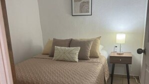 A bedroom with a bed, pillows, a nightstand with a lamp, and a framed picture on the wall. - Charming 2-bedroom house in Santa Ana with WiFi and AC (Santa Ana)