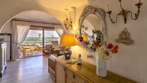 A room with a wooden cabinet, a mirror, a lamp, and a vase with flowers. There is a view of the ocean through an arched doorway.