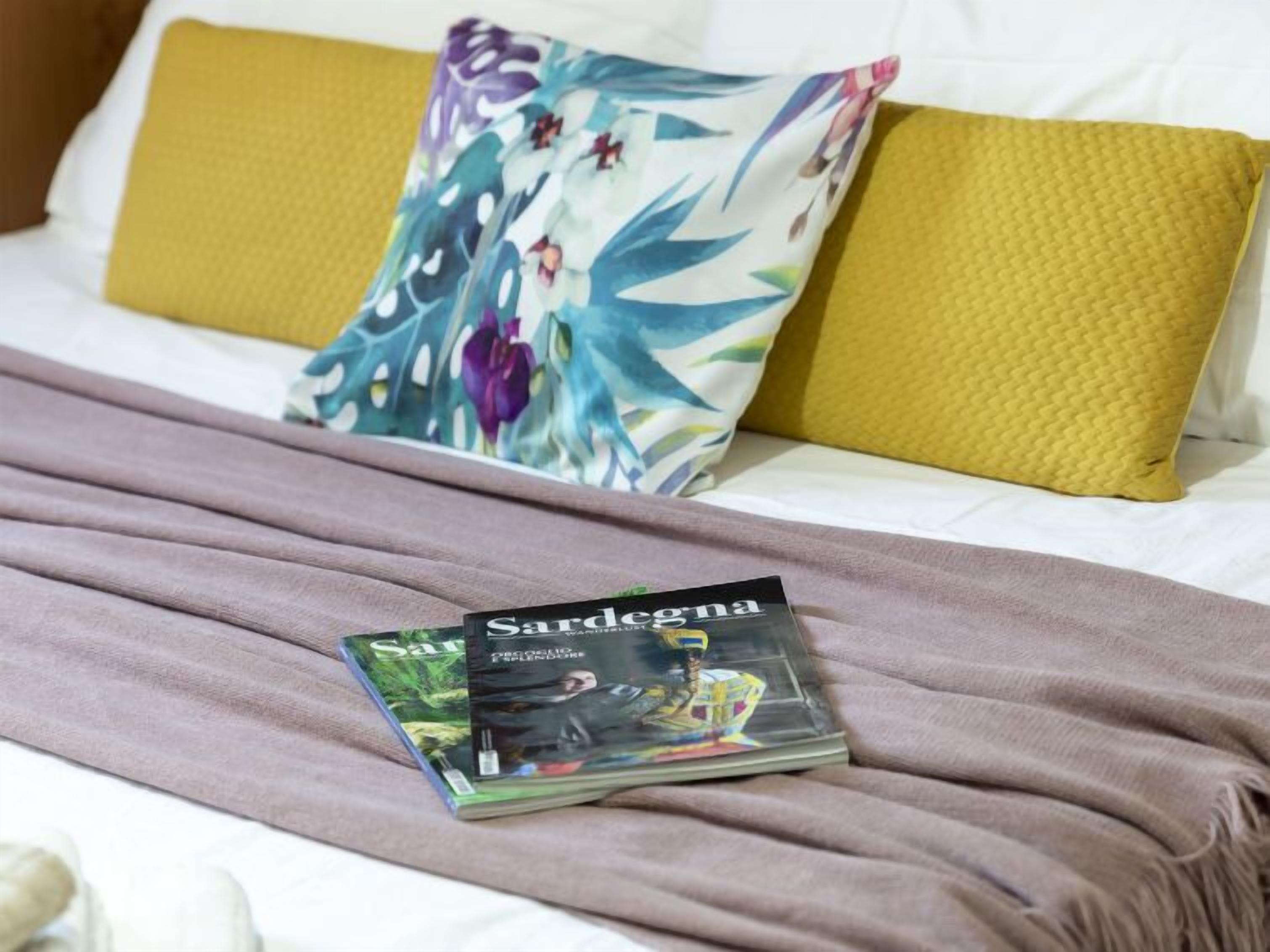 A bed with a white sheet, two pillows (one with a floral pattern, one solid yellow), and a pink blanket. Two books titled 'Sardegna' are placed on the bed.