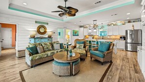 A spacious living room with wicker furniture, a ceiling fan, and a kitchen area with modern appliances. - Beach Retreat | Views, Bar, BBQ, Arcade, Games, Fenced Yard, Deck, Steps to Sand (Port Bolivar)