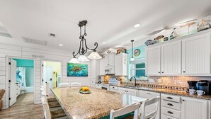 A spacious kitchen with white cabinetry, a granite island, and a dining area with white chairs. - Beach Retreat | Views, Bar, BBQ, Arcade, Games, Fenced Yard, Deck, Steps to Sand (Port Bolivar)