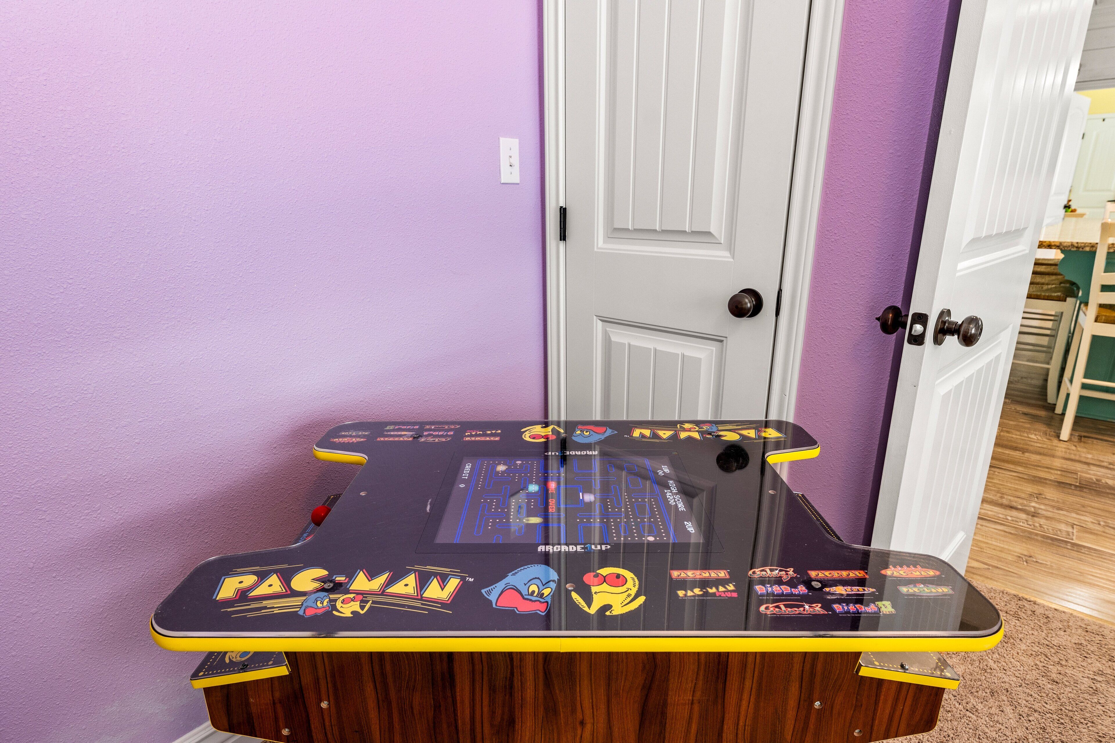 Une table d’arcade avec le jeu Pac-Man, dans une pièce aux murs violets et à la porte blanche.
