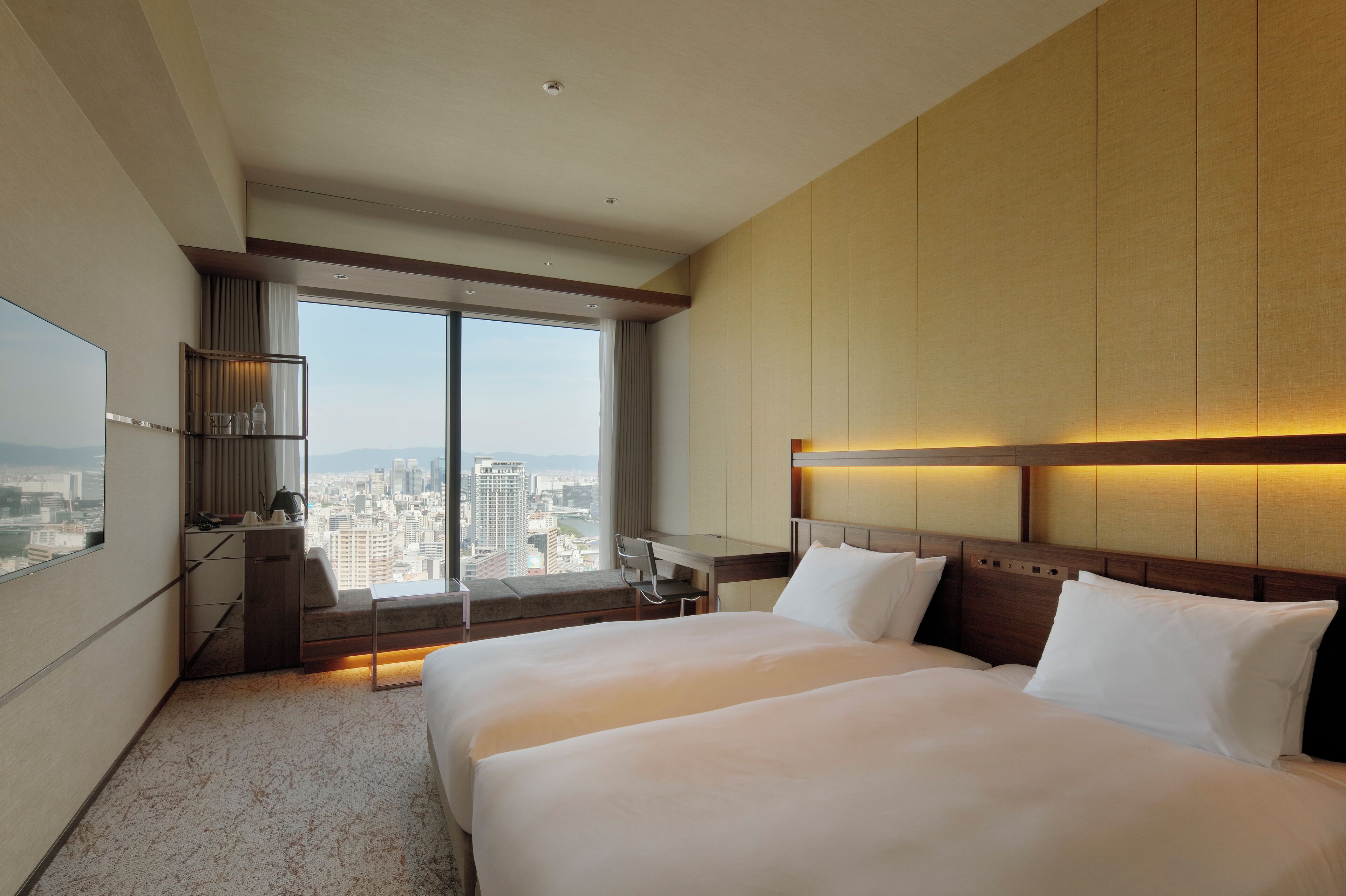 A hotel room with two beds, a desk, a chair, and a large window offering a city view.