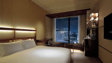 A hotel room with a large bed, a desk, a chair, a TV, and a window with a city view.