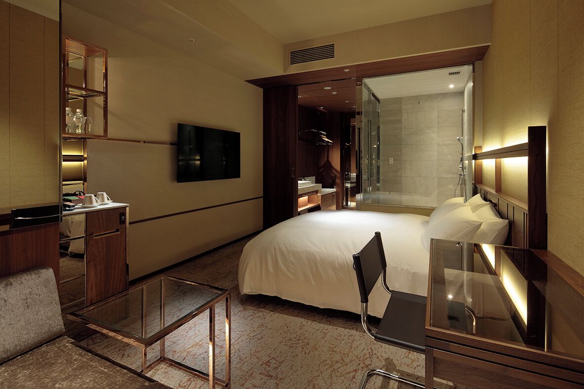 A modern hotel room with a large bed, a desk, a chair, a TV, a mirror, a wardrobe, and a shower area.