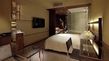 A modern hotel room with a large bed, a desk, a chair, a TV, a mirror, a wardrobe, and a shower area.