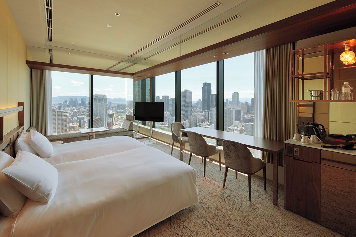 A hotel room with two beds, a desk, a TV, and a city view.