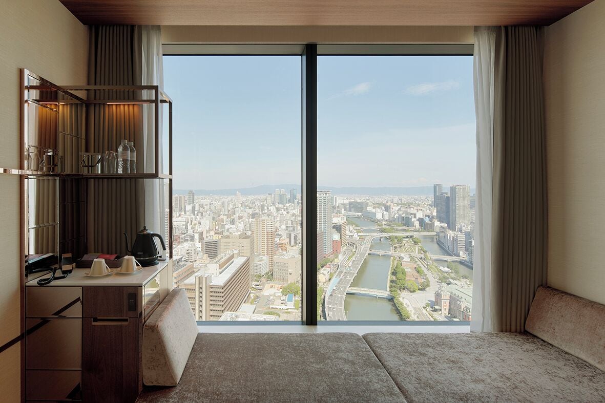 A hotel room with a large window offering a city view, a desk with a kettle, and a seating area.