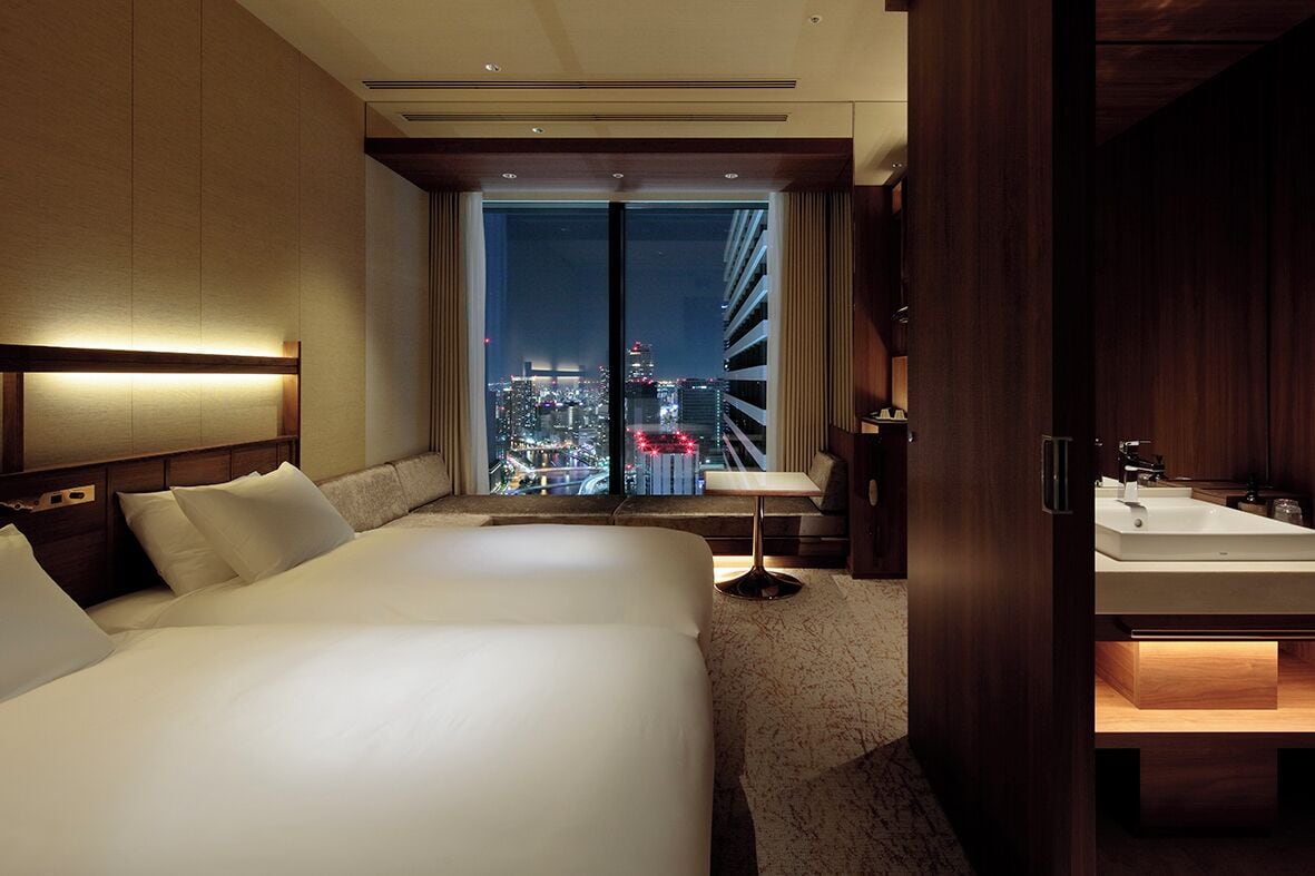 A hotel room with a large bed, a desk, and a view of a cityscape at night.