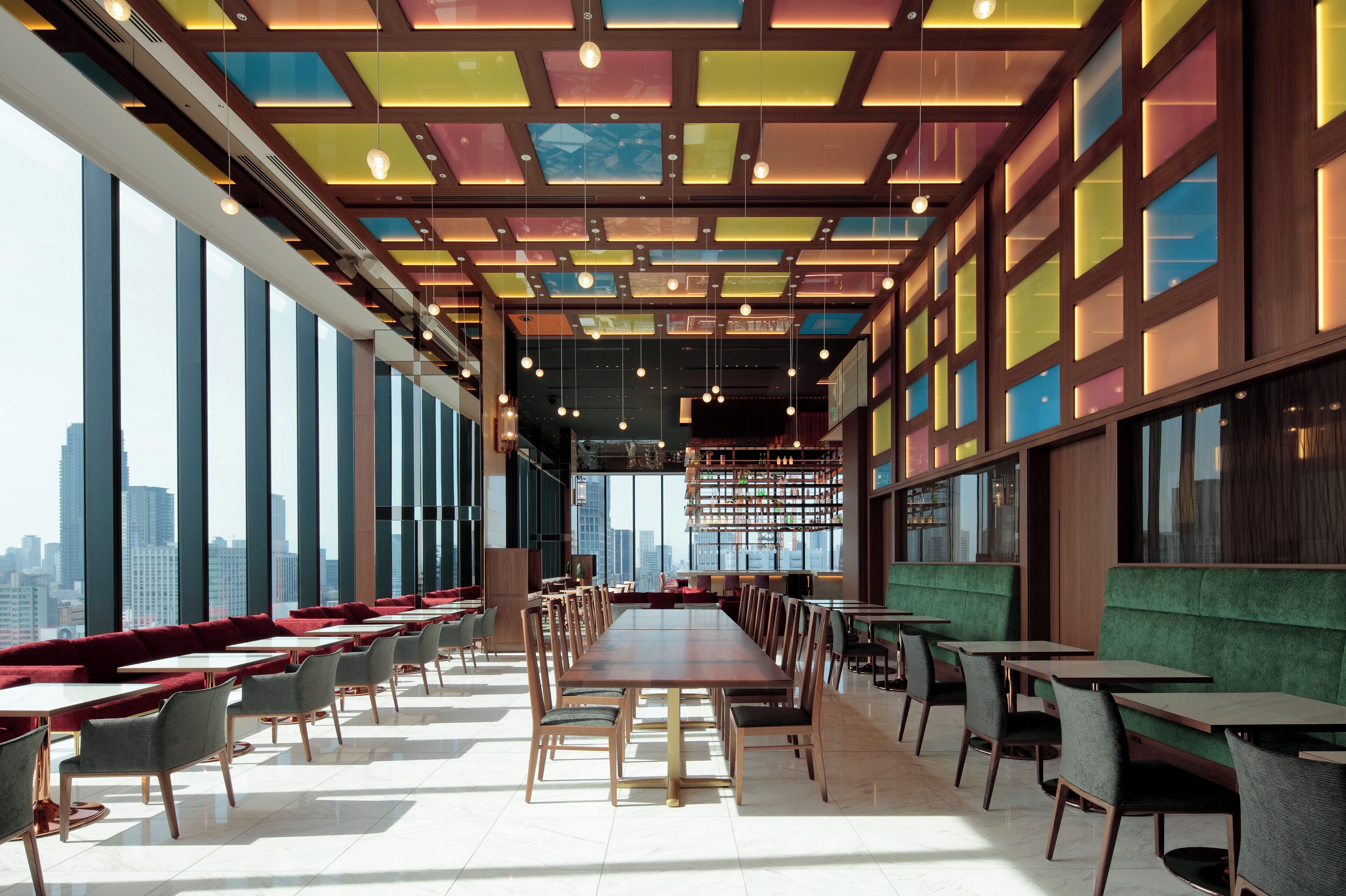 A modern dining area with long tables and chairs, large windows offering city views, and a colorful ceiling design.