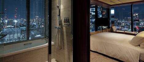 A modern hotel room with a large window offering a city view, a walk-in shower, and a bed with a cityscape view.