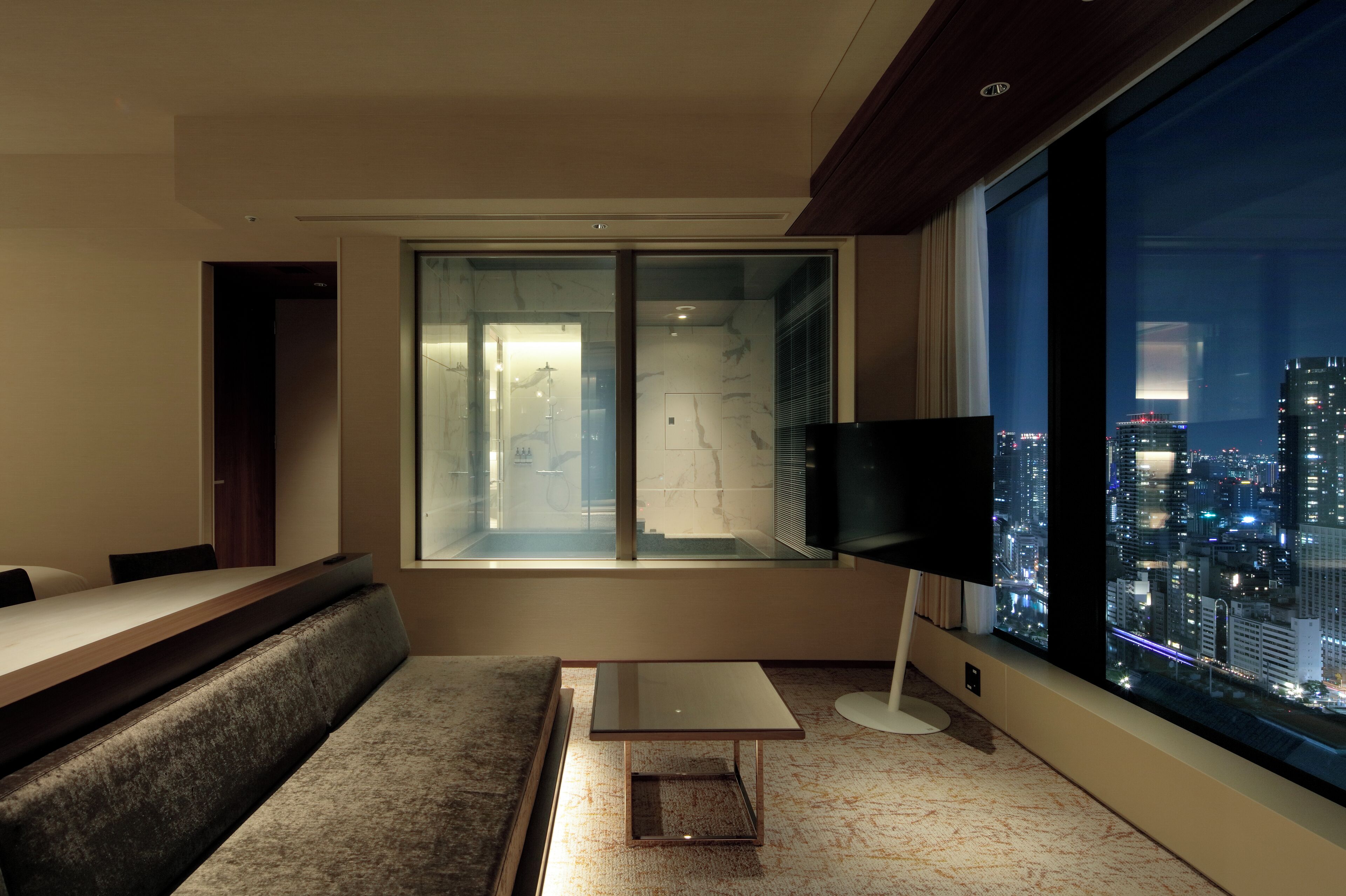 A modern hotel room with a large window offering a city view, a comfortable sofa, a small table, and a flat-screen TV.