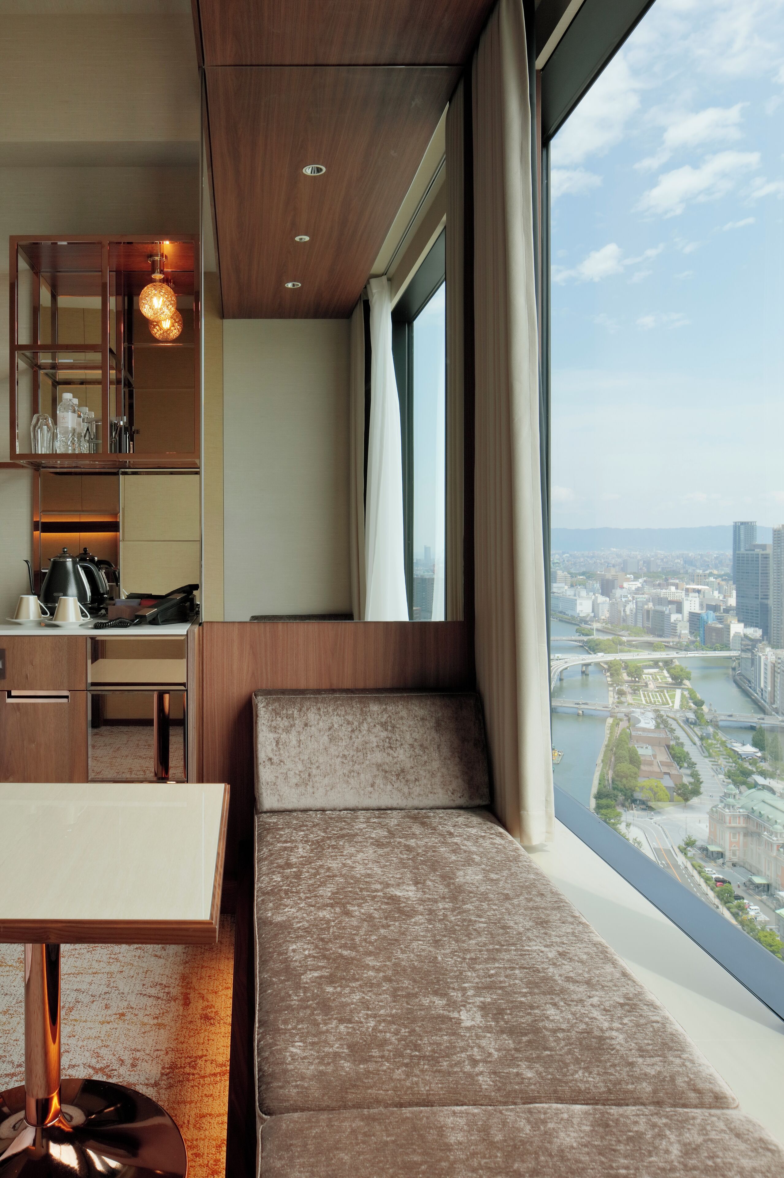 A modern hotel room with a large window, a built-in bench, and a kitchenette.