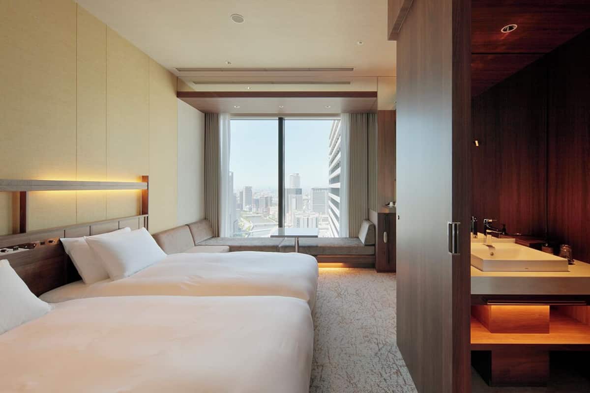 Une chambre d’hôtel moderne dotée d’un grand lit, offrant une vue sur la ville, d’une armoire en bois et d’une salle de bain avec un grand lavabo.