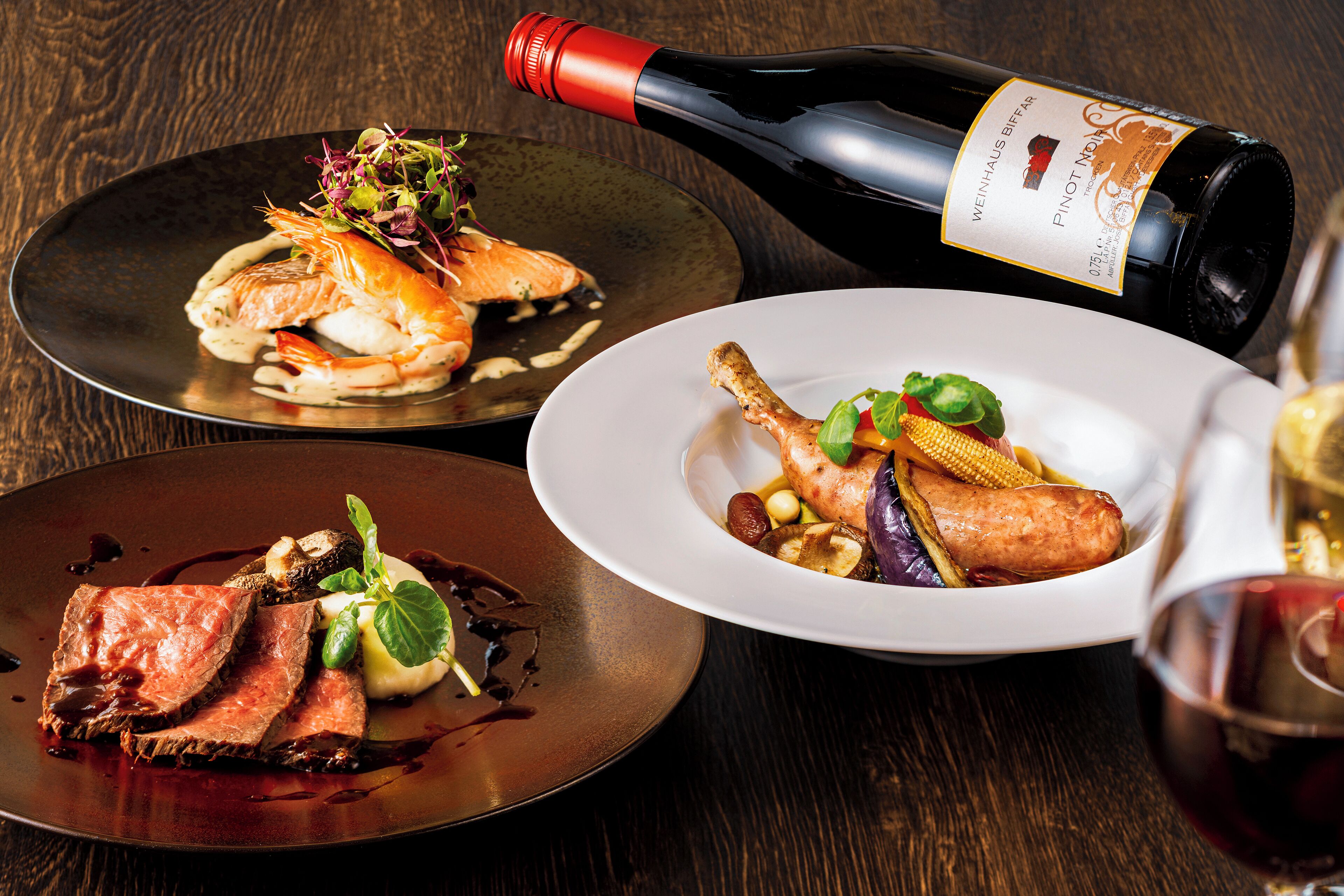 Three plates of food on a wooden table with a bottle of wine and a glass of wine.