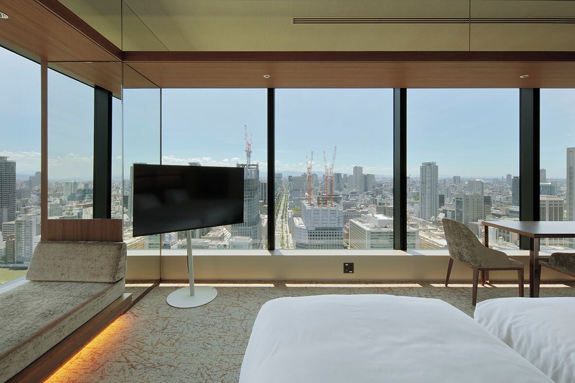 A modern hotel room with a large city view, a flat-screen TV, a bed, a chair, and a small table.