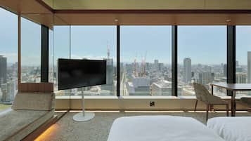 A modern hotel room with a large city view, a flat-screen TV, a bed, a chair, and a small table.