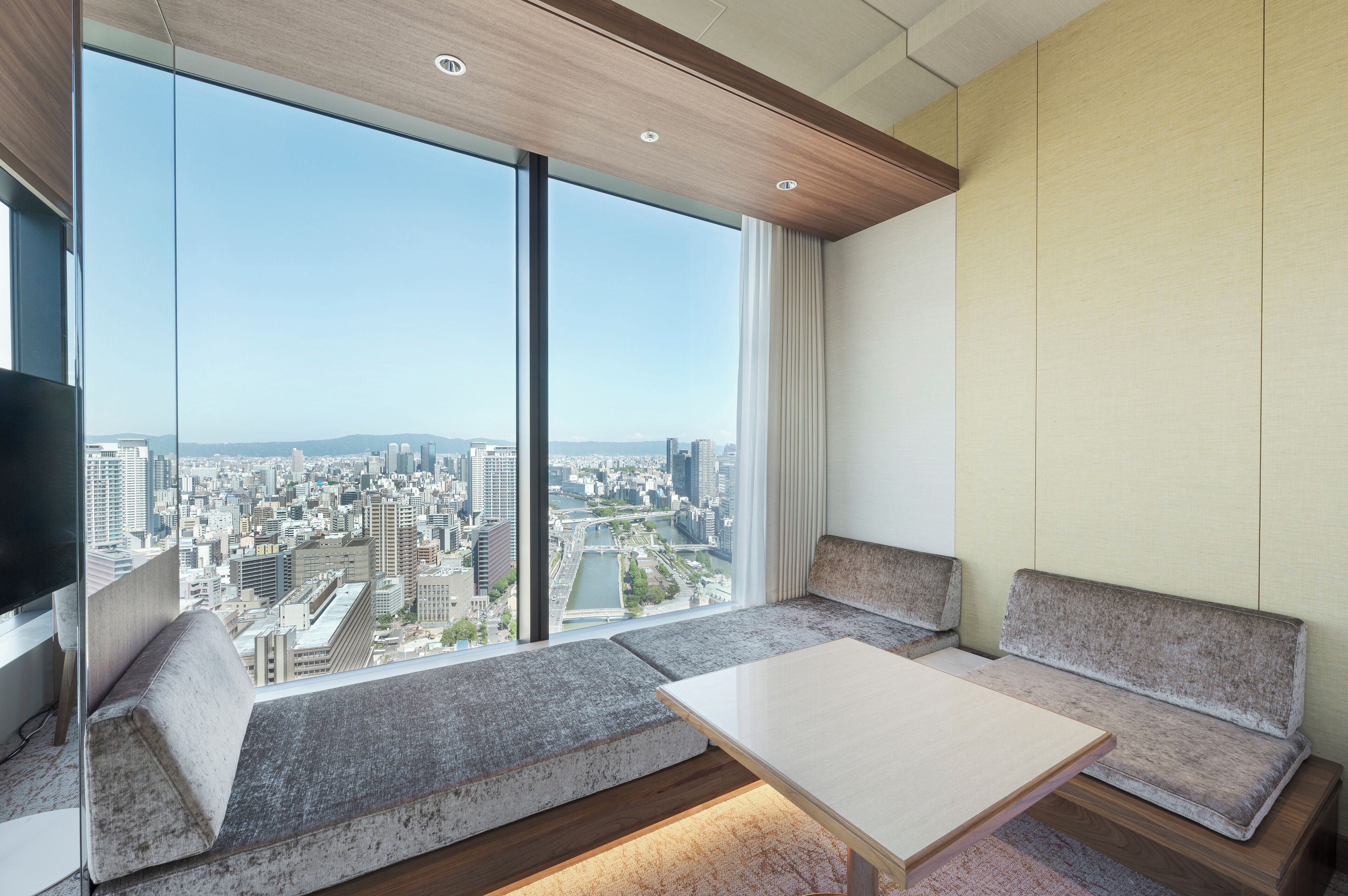A modern hotel room with a large window offering a city view, a flat-screen TV, and a sofa with a table.