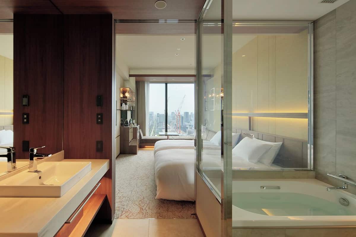 Une chambre d’hôtel moderne dotée d’un grand lit, d’une salle de bain séparée avec baignoire et offrant une vue sur la ville grâce à de grandes fenêtres.
