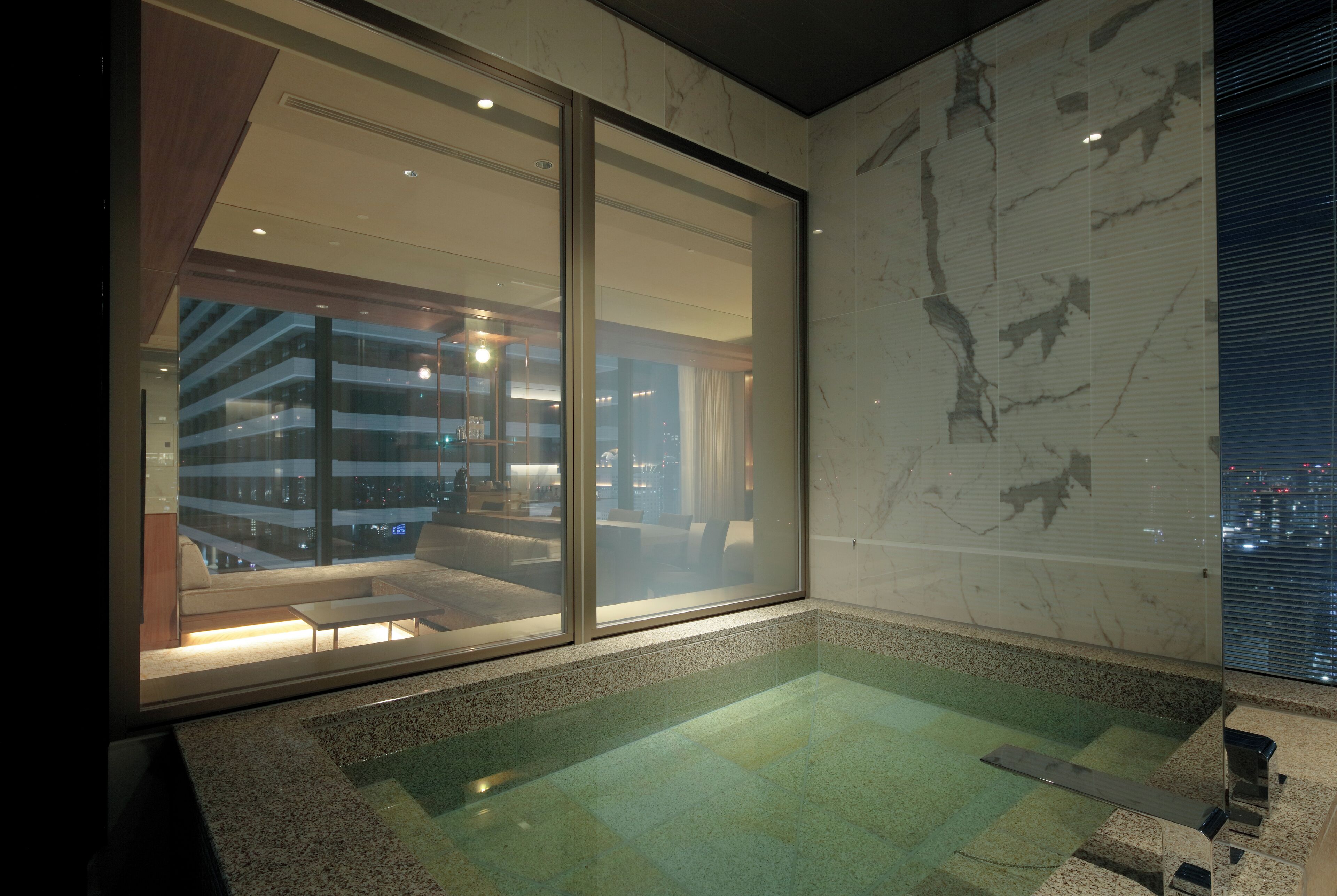 A modern hotel room with a large sliding glass door leading to a balcony, a spa area with a hot tub, and a city view.