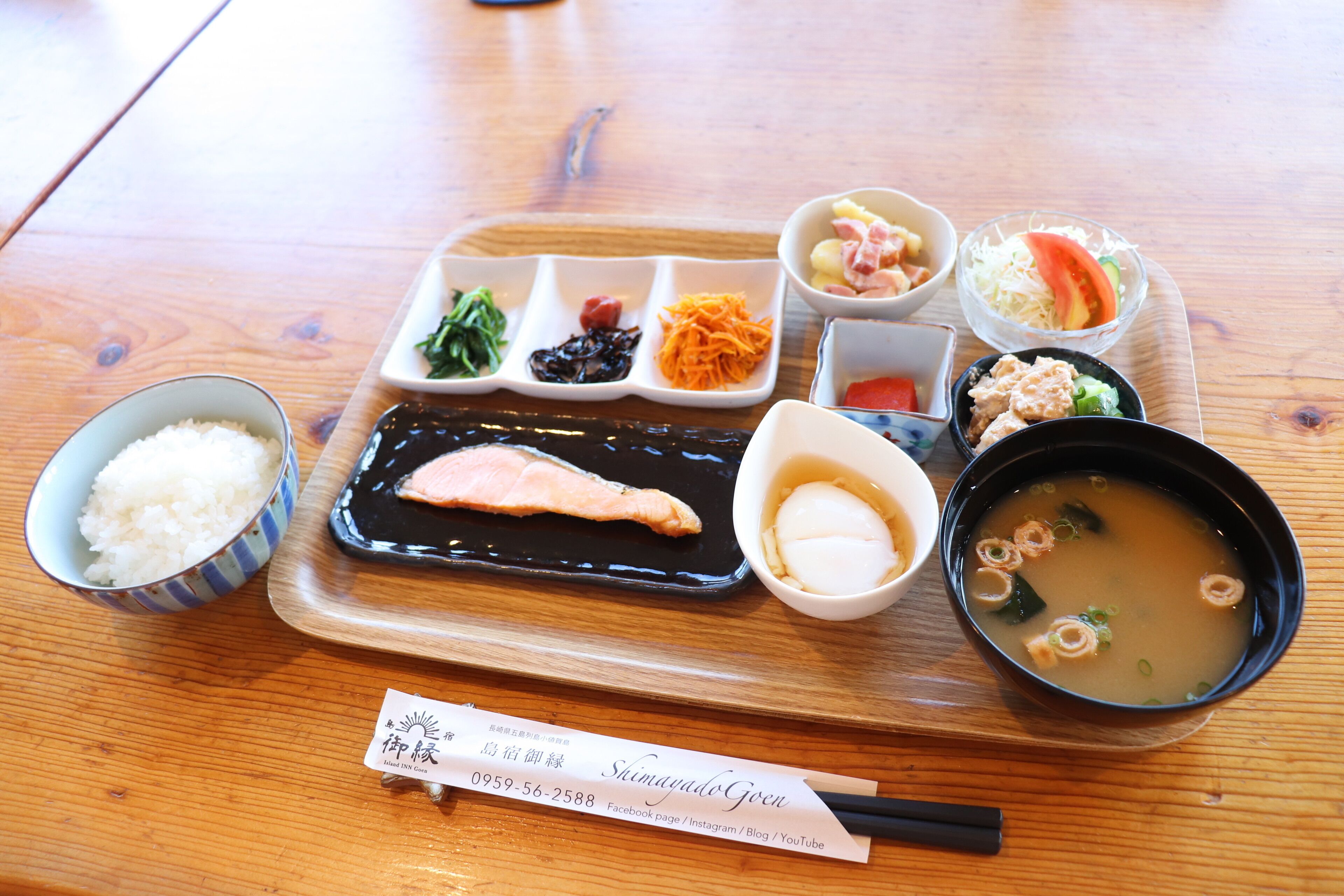 A wooden table with a meal tray containing rice, salmon, soup, and various side dishes.