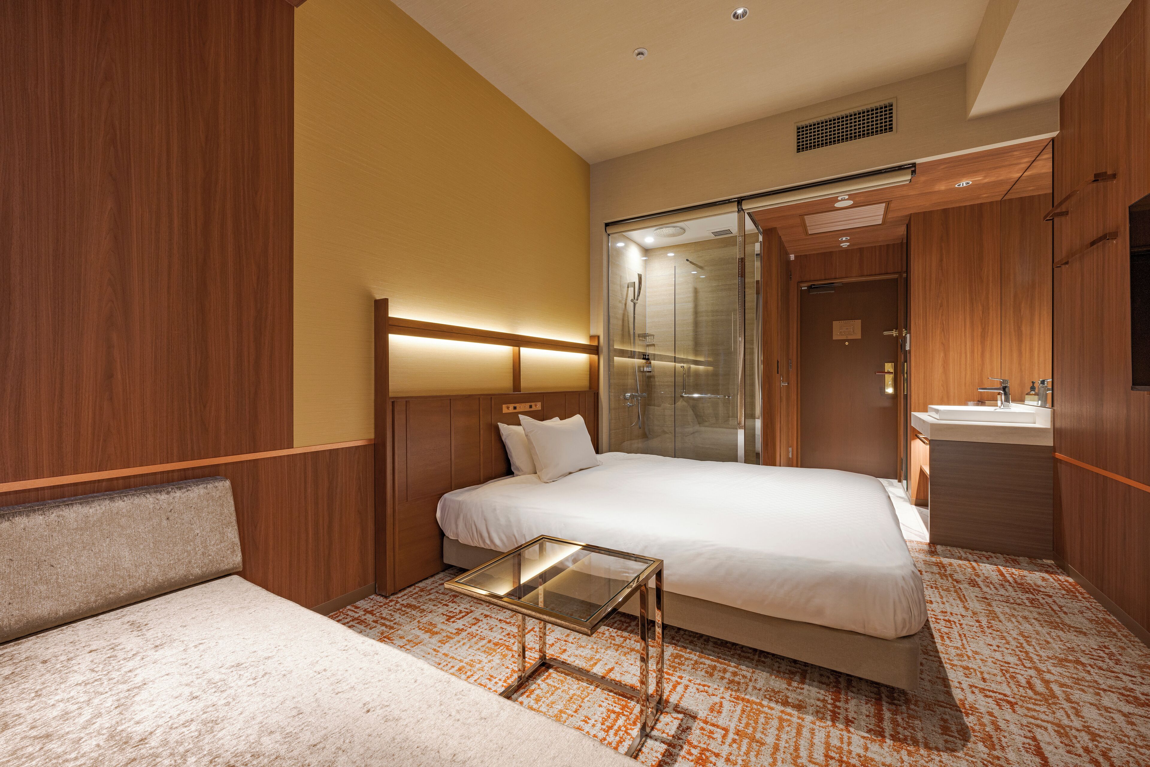 A hotel room with a bed, a glass table, a shower, and a sink.