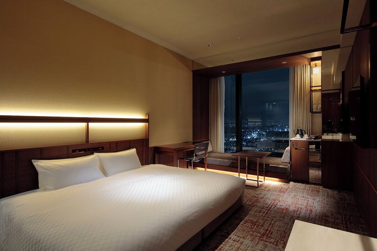 Een hotelkamer met een groot bed, een bureau, een stoel en uitzicht op de stad 's nachts.