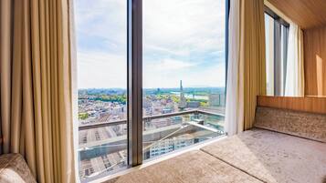 A high-rise room with a large window offering a city view, a cushioned bench, and a wooden ceiling.